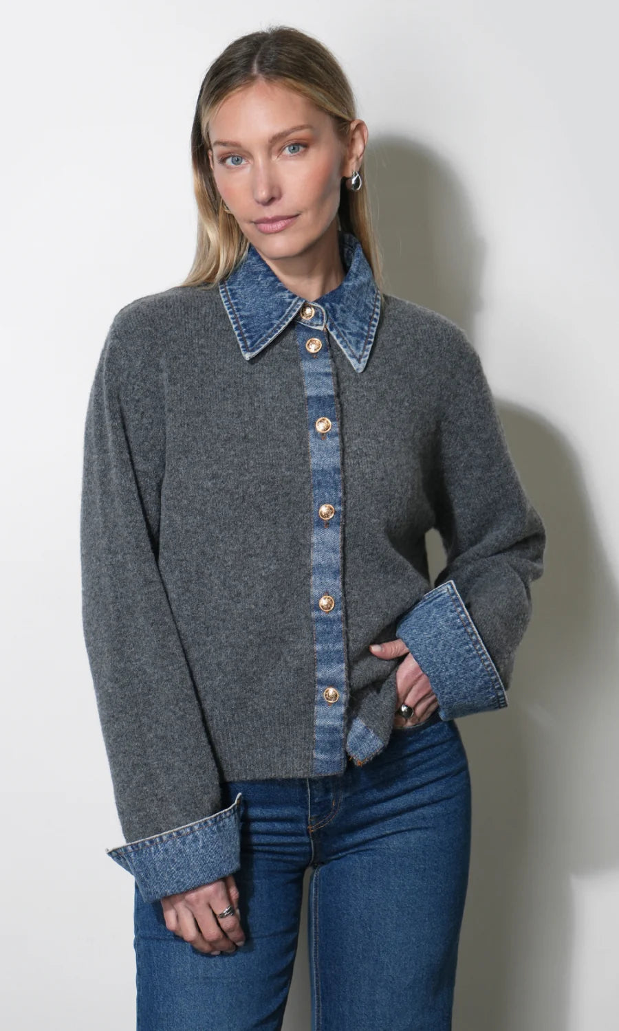 A woman modeling a gray knit cardigan with a blue denim collar, button placket, and cuffed sleeves, paired with blue jeans against a plain light gray wall.