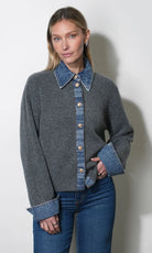 A woman modeling a gray knit cardigan with a blue denim collar, button placket, and cuffed sleeves, paired with blue jeans against a plain light gray wall.
