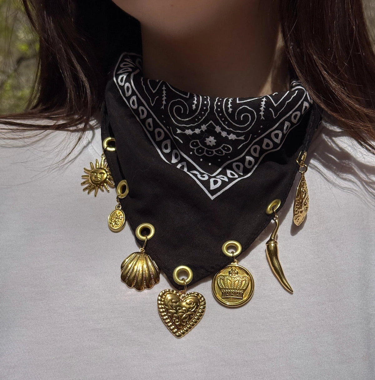 A close-up of a person wearing a black bandana-style necklace with white paisley print and multiple dangling gold-tone charms, including a heart, shell, sun, crown, and chili pepper shapes.
