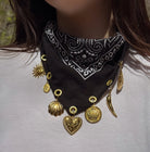 A close-up of a person wearing a black bandana-style necklace with white paisley print and multiple dangling gold-tone charms, including a heart, shell, sun, crown, and chili pepper shapes.