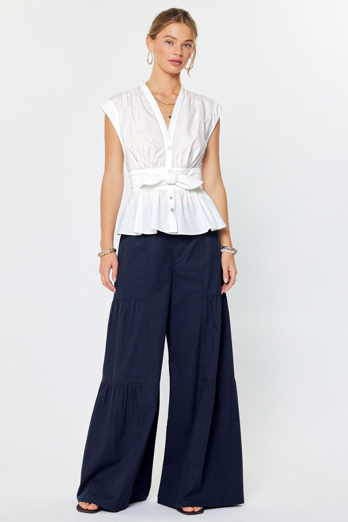 Woman wearing a white sleeveless top with a belt and navy pants on a white background
