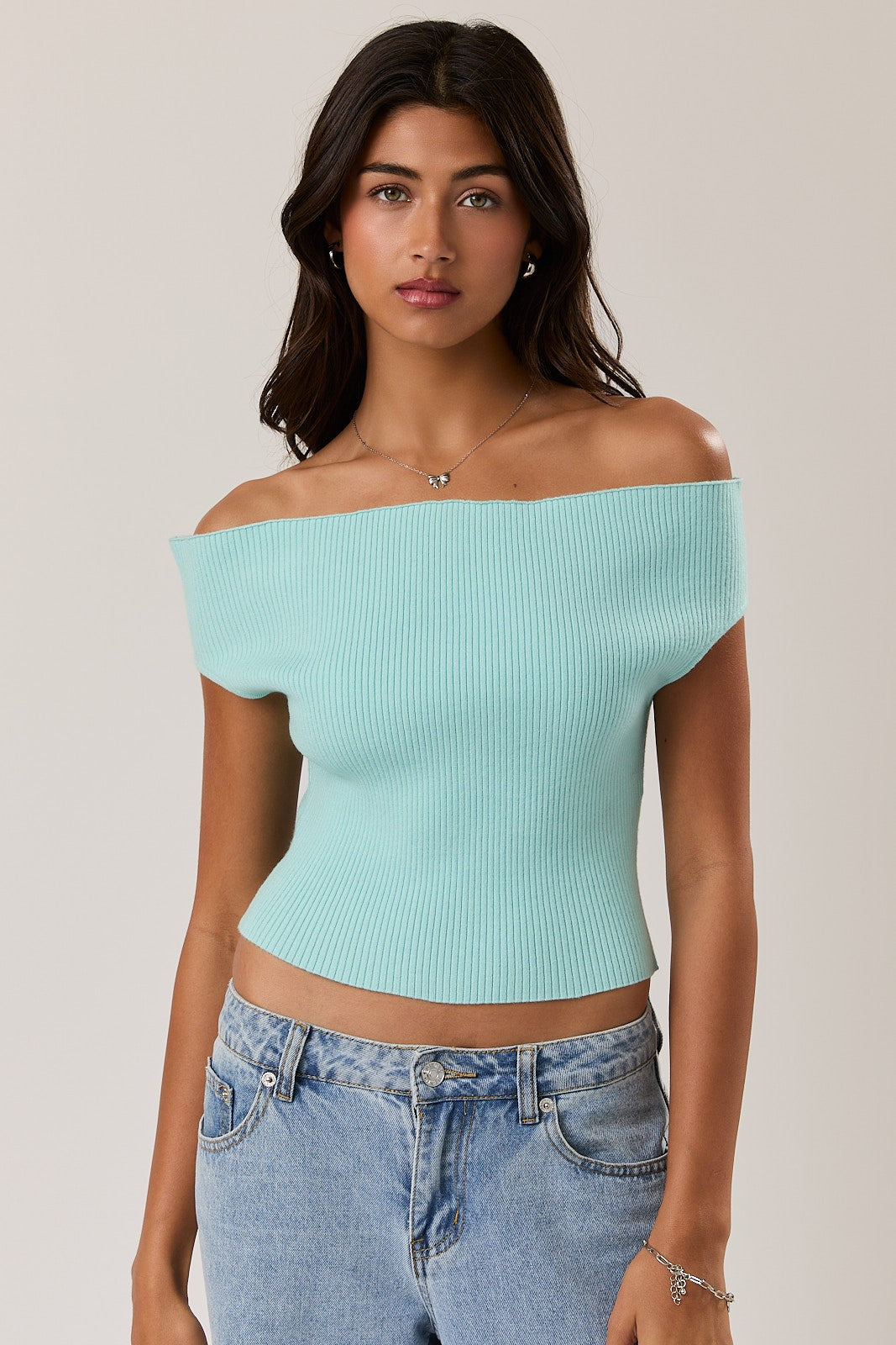 Woman wearing a light blue off-shoulder top and blue jeans against a plain background