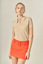 Woman wearing a beige polo shirt and red skirt against a neutral background