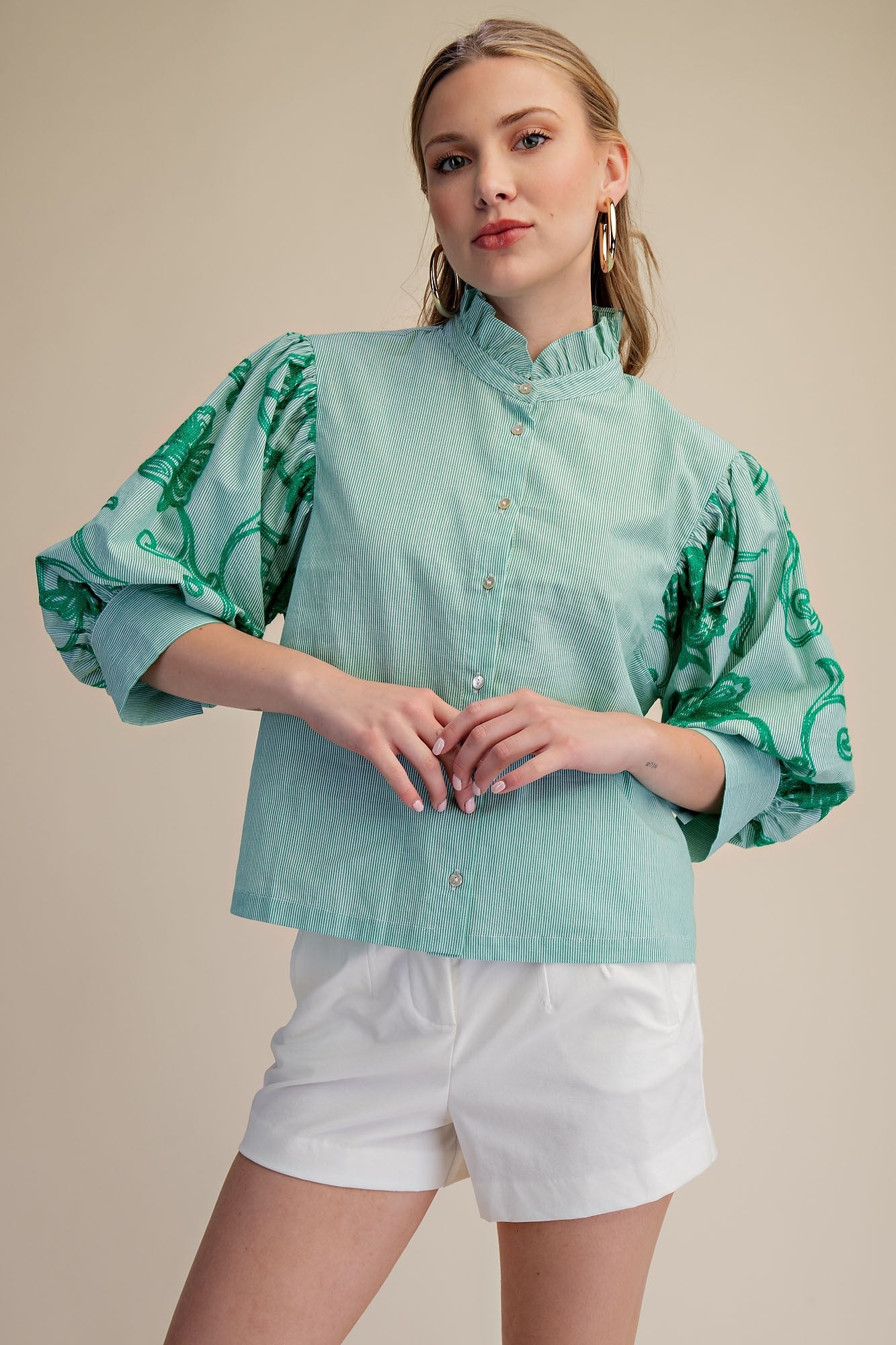 Woman wearing a light green blouse with floral patterns and white shorts on a beige background