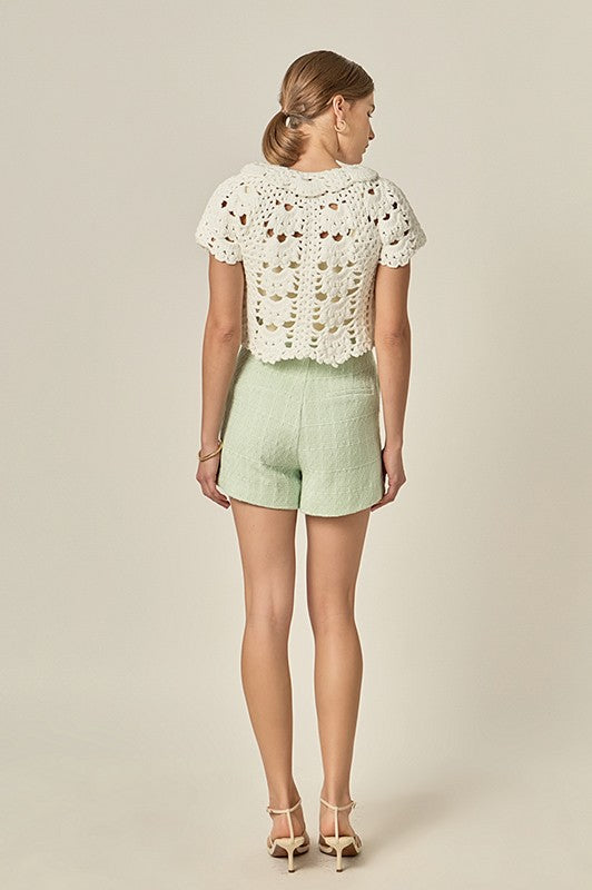 Woman wearing a white lace top and light green shorts on a beige background