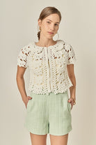 Woman wearing a white lace top and light green shorts on a beige background