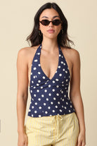 Woman wearing a navy blue halter top with white polka dots and yellow pants against a beige background