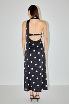 A woman viewed from the back wearing a black maxi dress with a white polka dot pattern and an open back cutout, paired with black strappy heels against a plain light gray studio background.