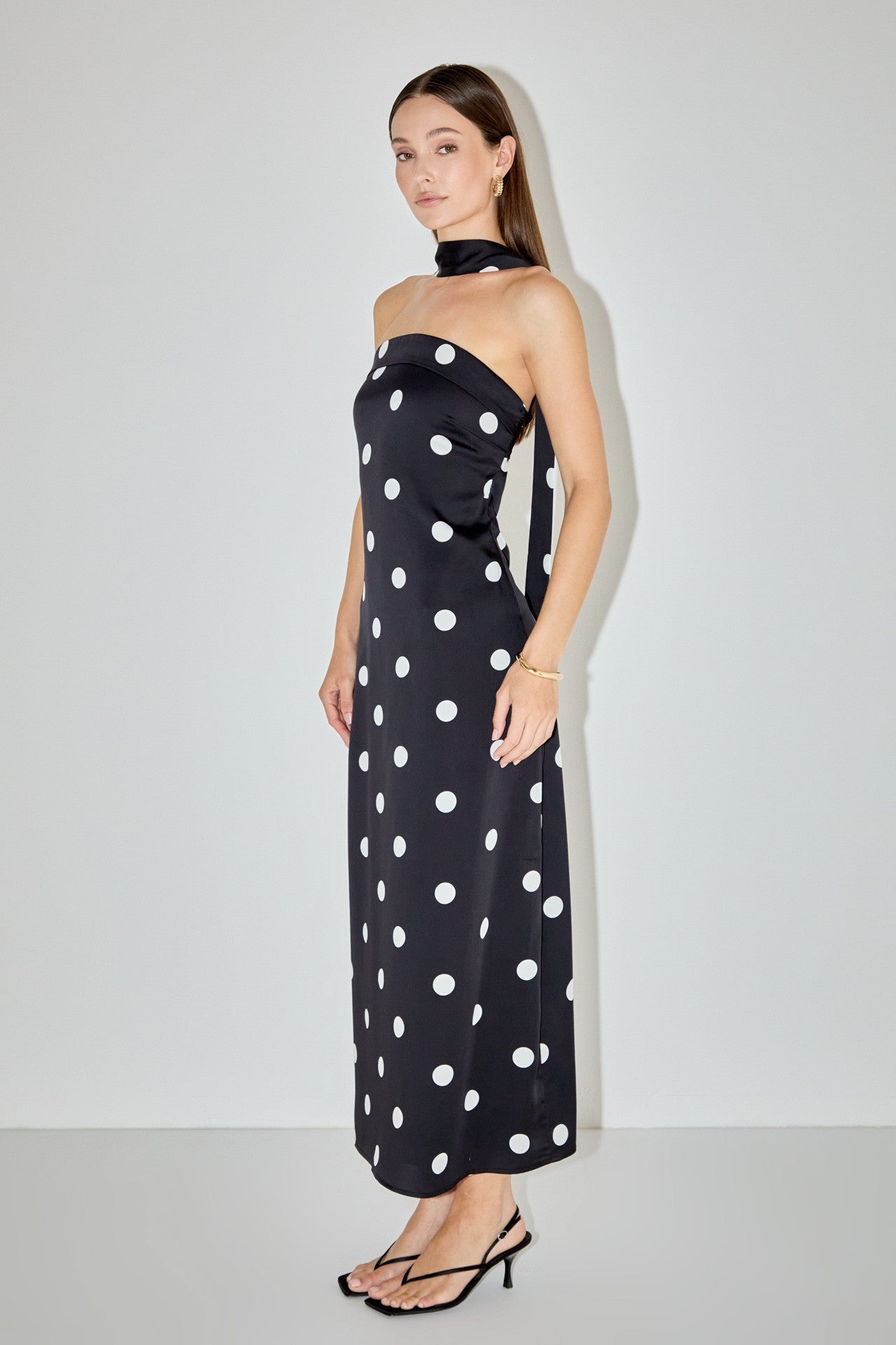 A woman modeling a strapless black maxi dress with large white polka dots and a matching black halter-style neck band, standing against a plain light gray studio background.