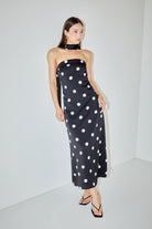 A woman modeling a strapless black midi dress with large white polka dots, paired with a matching black neck scarf and black strappy sandals, standing in a minimalist white studio corner.