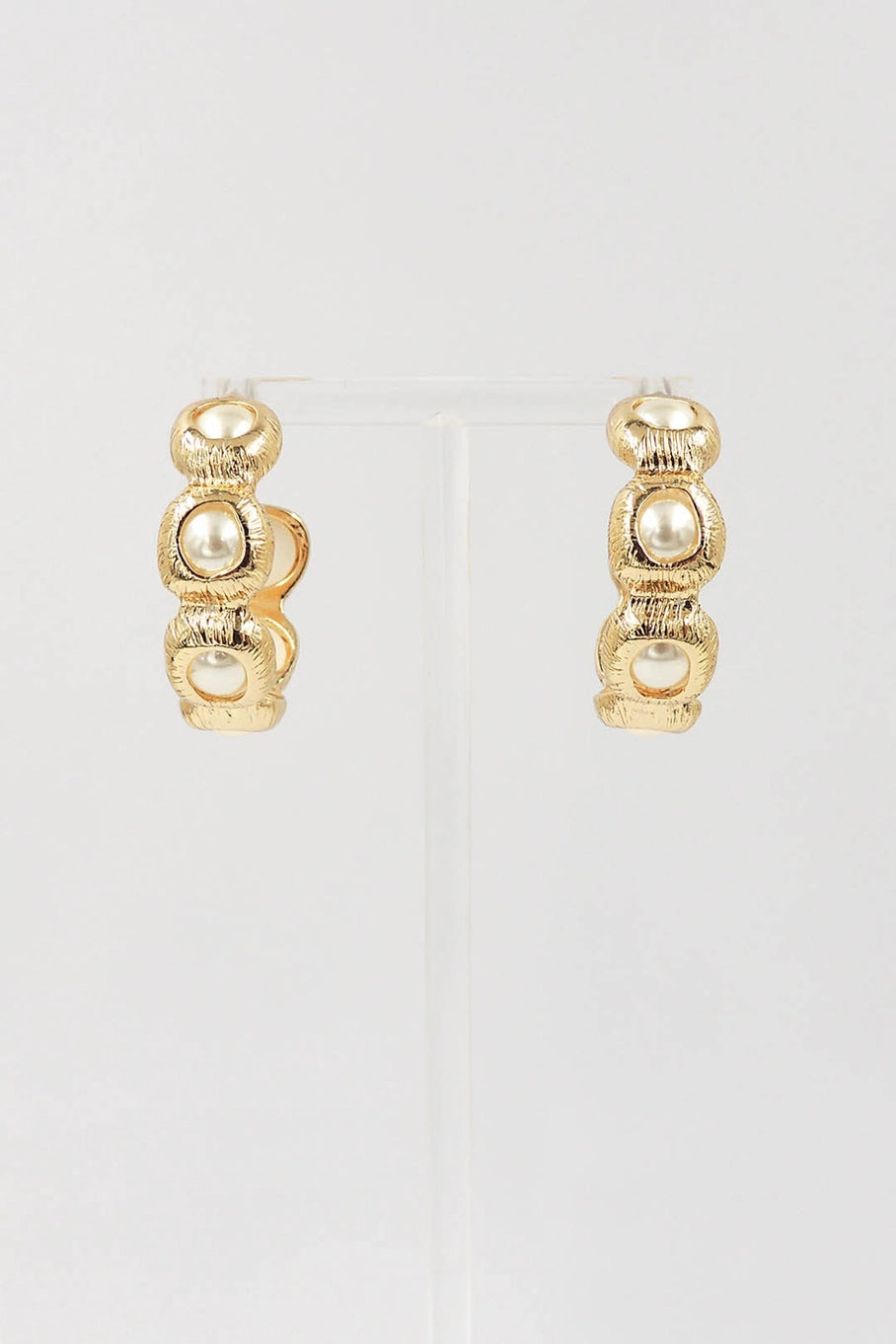 A pair of gold huggie hoop earrings with three stacked textured gold beads and a central white pearl, displayed on a clear stand against a light gray background.