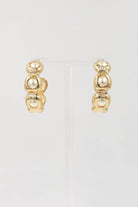 A pair of gold huggie hoop earrings with three stacked textured gold beads and a central white pearl, displayed on a clear stand against a light gray background.