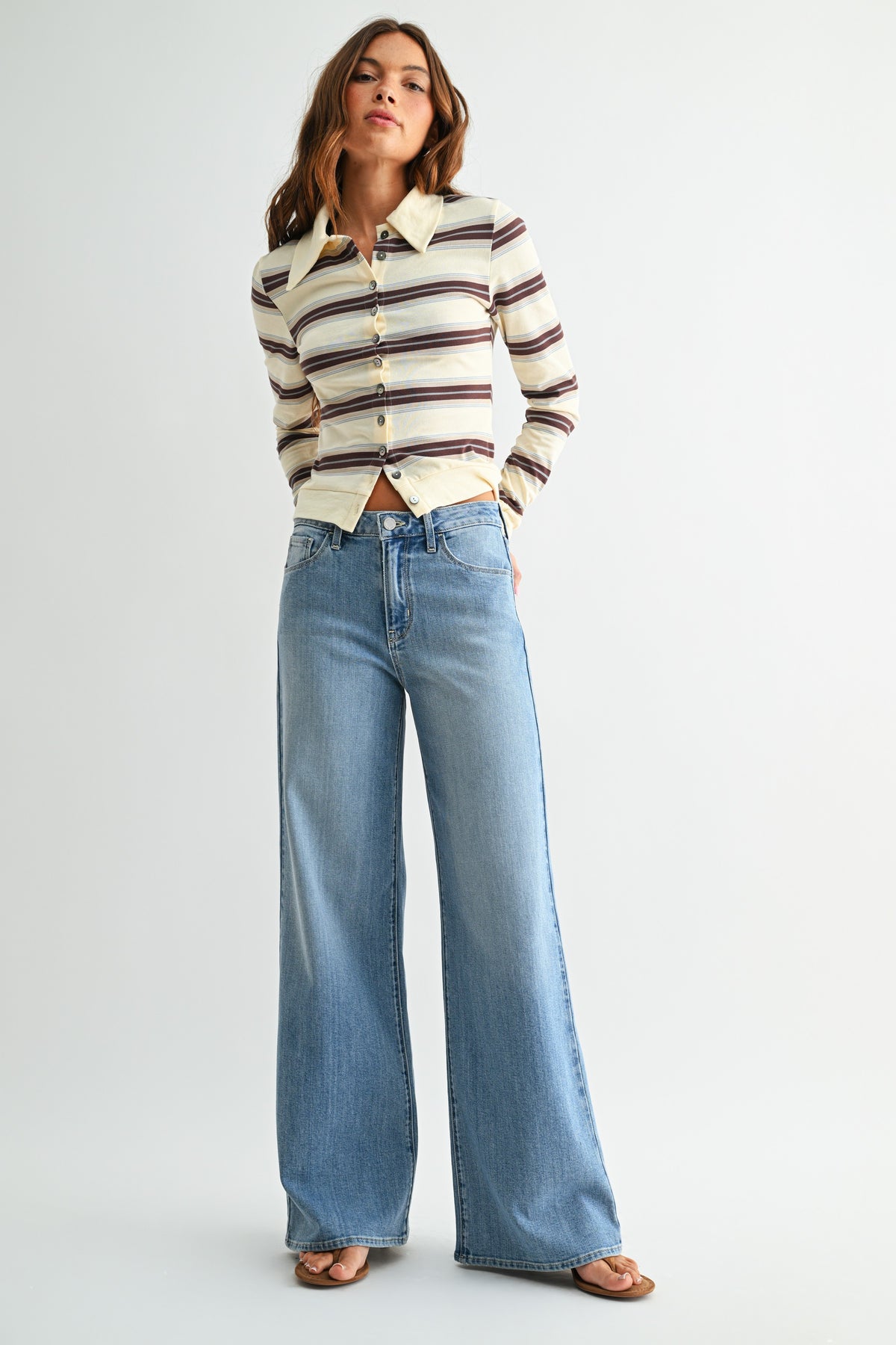 A woman modeling a cream long-sleeve button-up shirt with dark brown and burgundy horizontal stripes, paired with high-waisted, wide-leg light blue jeans and brown flat sandals, against a plain light gray studio background.