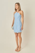 A woman modeling a light blue strapless mini dress with a scalloped edge and white piping details, paired with white strappy high-heeled sandals and holding a small clutch, set against a plain beige studio background.