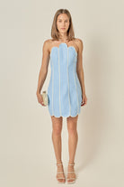 A woman modeling a strapless light blue dress with vertical pleated panels and white piping, standing against a plain beige studio background.
