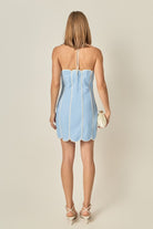 A woman shown from the back wearing a light blue strapless mini dress with white trim and a scalloped hem, holding a small cream clutch against a plain beige studio background.