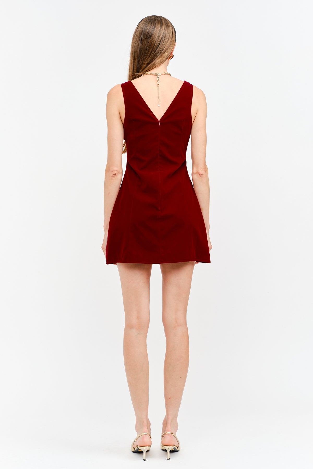 A woman viewed from the back wearing a sleeveless deep red mini dress with a V-neckline and flared skirt, paired with metallic high-heeled sandals against a plain white studio background.