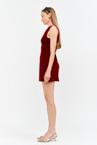 A woman in profile wearing a sleeveless deep red velvet mini dress and gold pointed-toe slingback heels, standing against a plain white studio background.