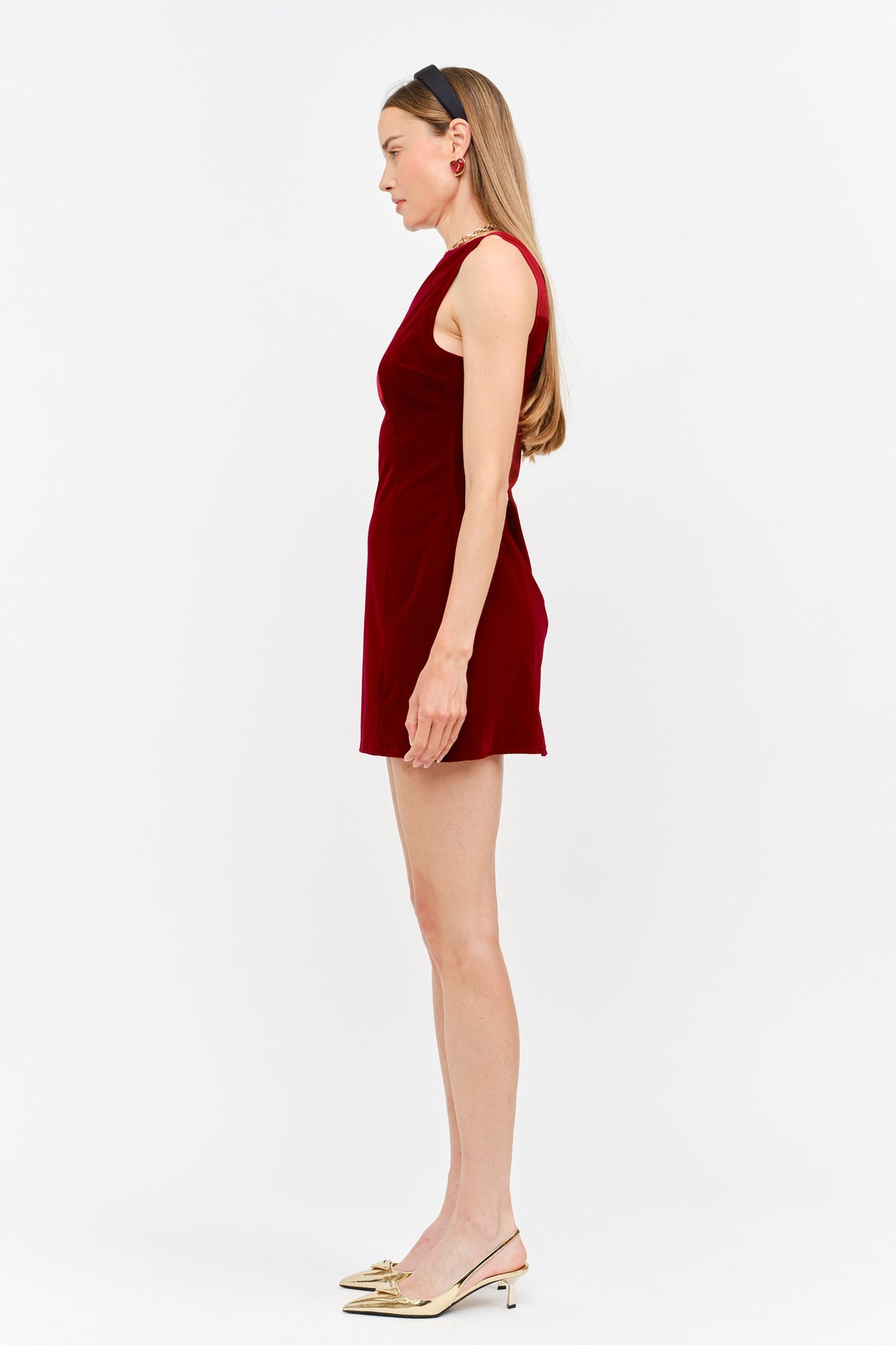 A woman in profile wearing a sleeveless deep red velvet mini dress and gold pointed-toe slingback heels, standing against a plain white studio background.