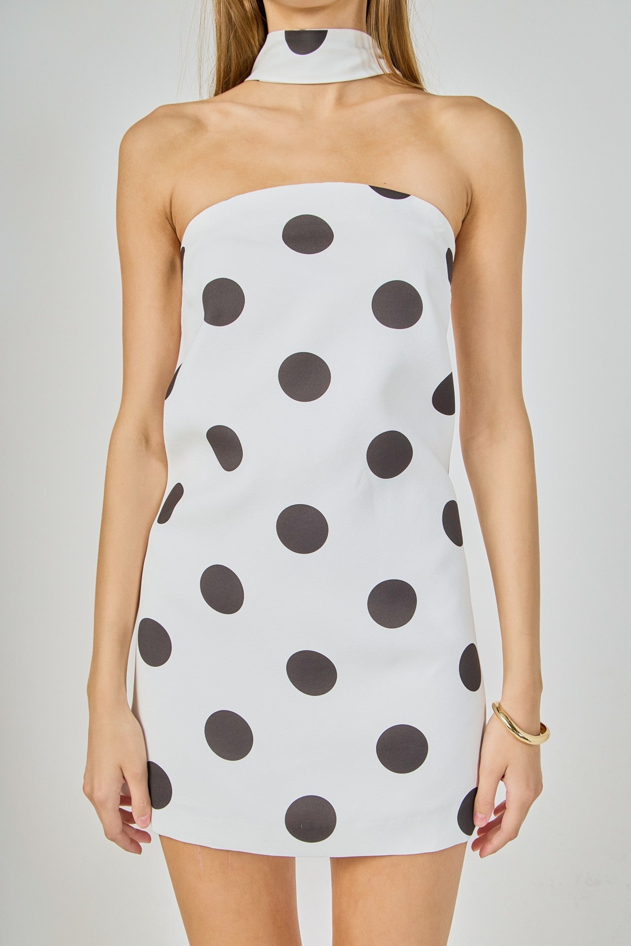 A woman wearing a strapless halter-neck mini dress with a white base and large dark gray polka dots, shown against a light gray studio background.