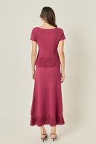 A woman viewed from the back wearing a magenta maxi dress with short sleeves, a crew neckline, and fringe detailing at the waist and hem, paired with black high-heeled sandals against a light beige studio background.