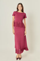 A woman modeling a dusty rose pink knit dress with short sleeves, a boat neckline, and fringe detailing at the peplum and hem, paired with dark brown pointed-toe heels against a light gray studio background.