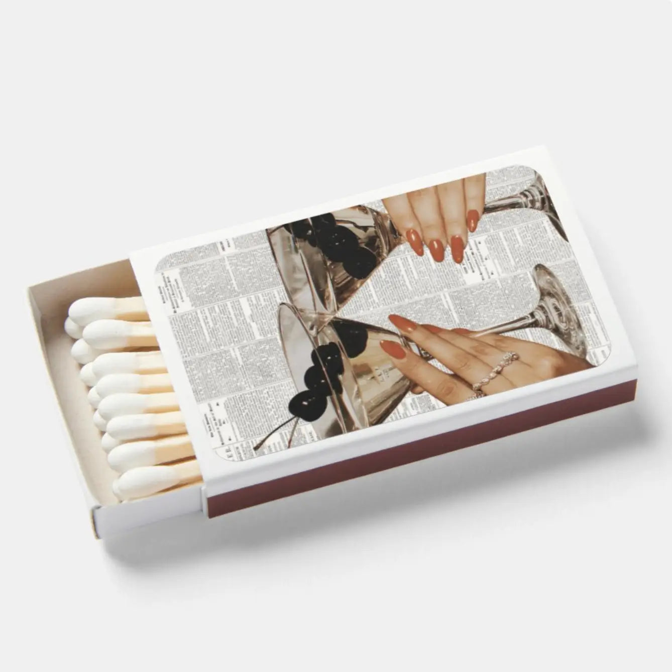 A matchbox with white matches visible on the left, photographed on a clean light gray background. The top lid features a printed collage design with a woman's hands, red-orange manicured nails, rings, and a vintage-style typewriter over a newspaper page.