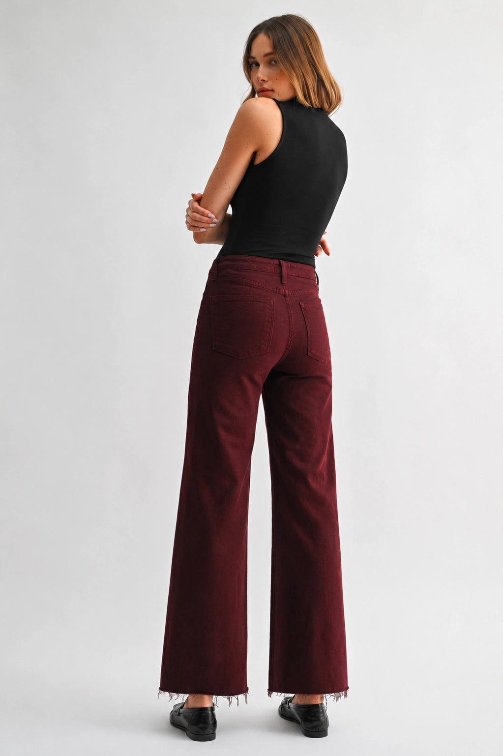 A woman shown from the back wearing high-waisted, wide-leg burgundy corduroy pants with a frayed hem, a black sleeveless top, and black shoes, against a light gray studio background.