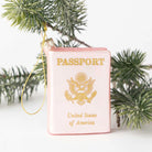 A novelty Christmas ornament shaped like a pink United States passport with gold foil lettering and emblem, hanging from a gold string and styled with green pine branches against a white background.