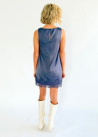 A woman viewed from behind wearing a sleeveless slate blue shift dress with a lace hem, paired with off-white cowboy boots, against a plain white studio background.