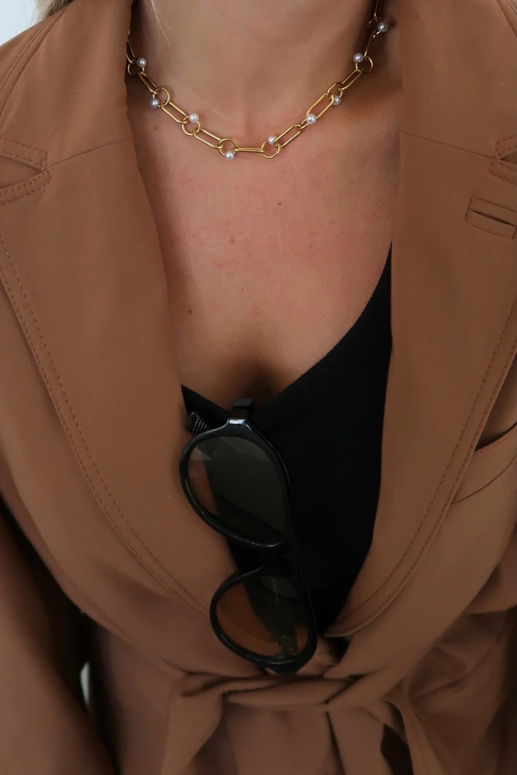 Close-up of a person wearing a brown coat with a gold chain necklace and sunglasses hanging from the collar.