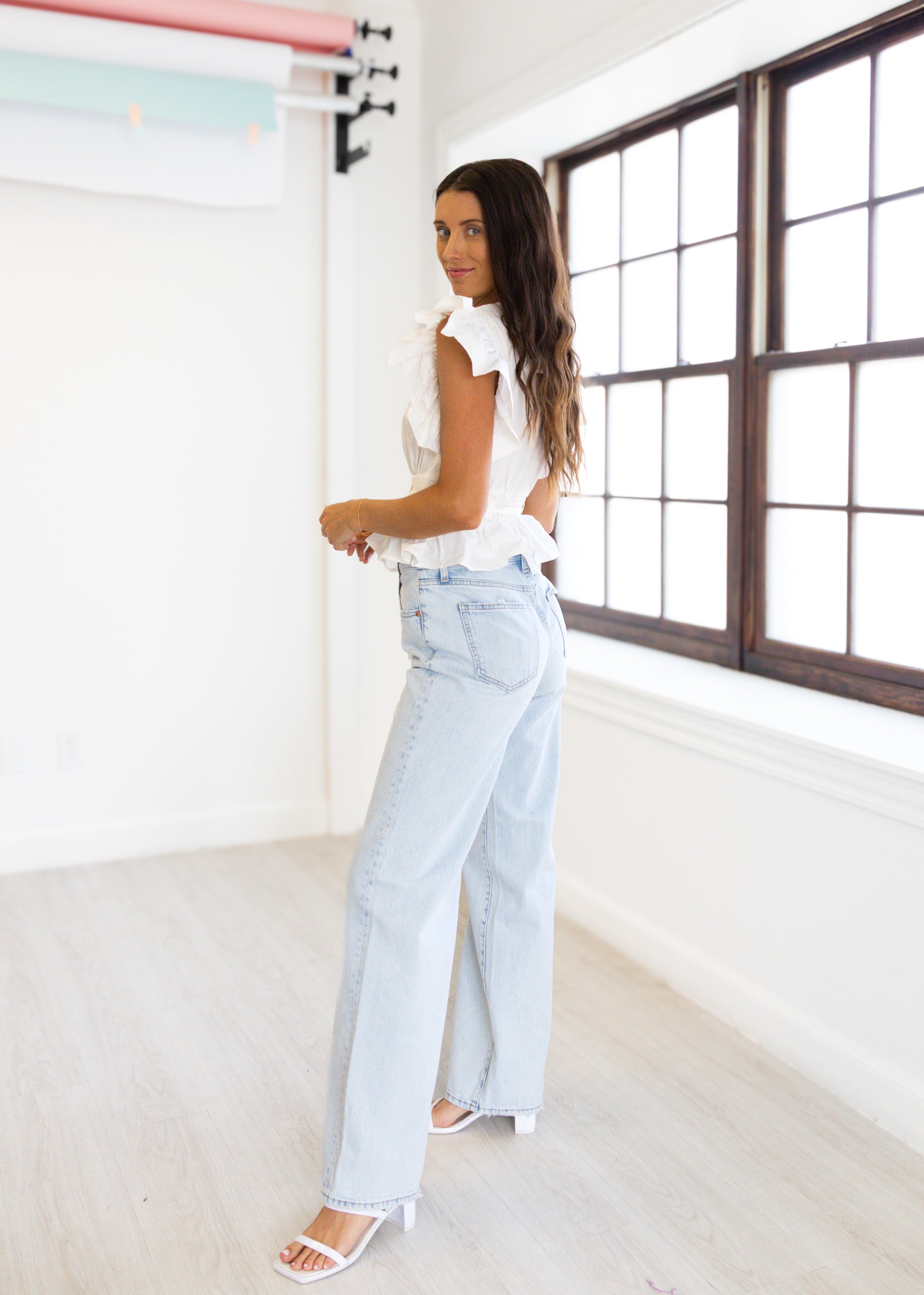 Woman in a white top and light blue jeans standing in a bright room with large windows.