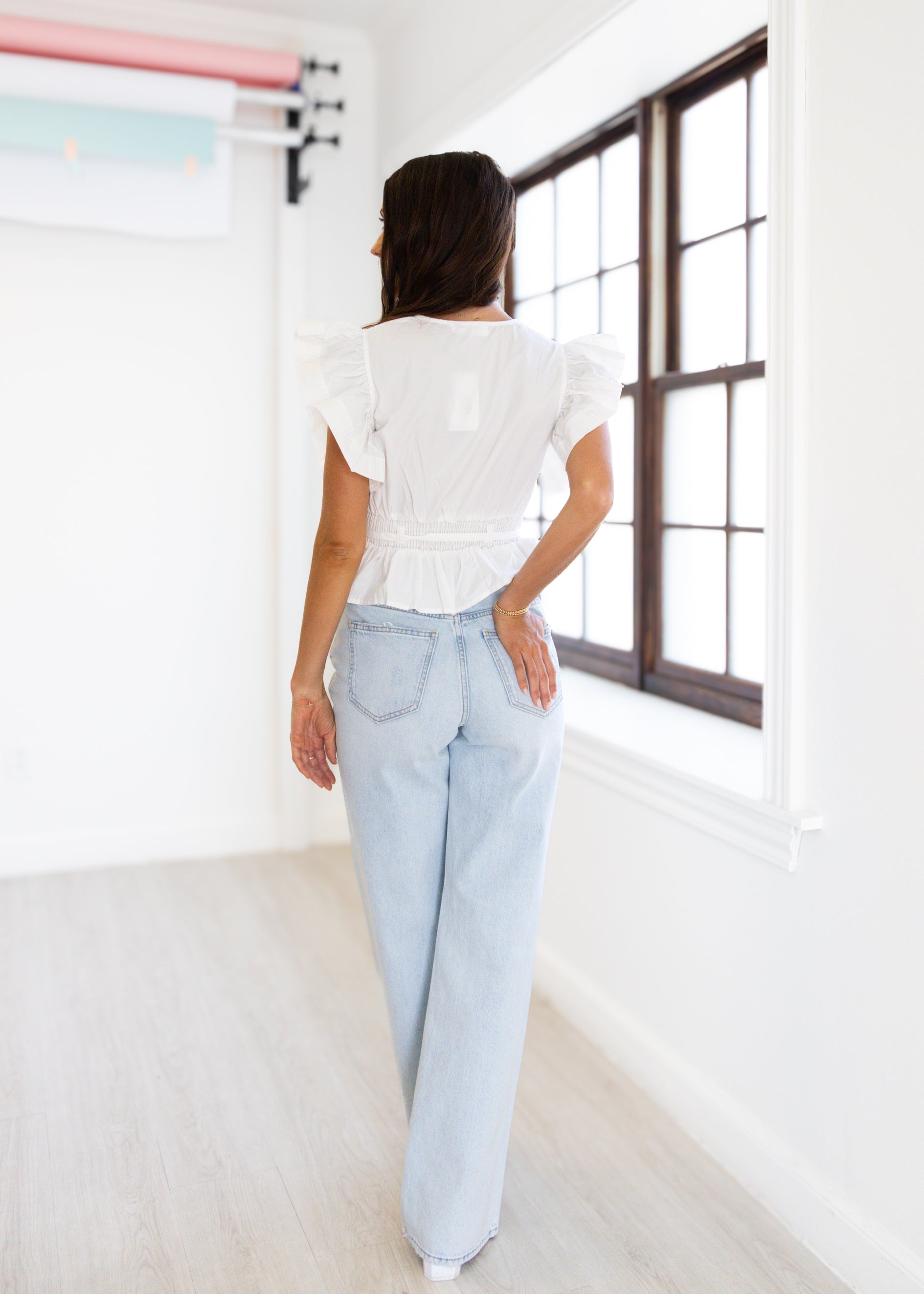 Person wearing a white top and light blue jeans in a bright room with large windows.