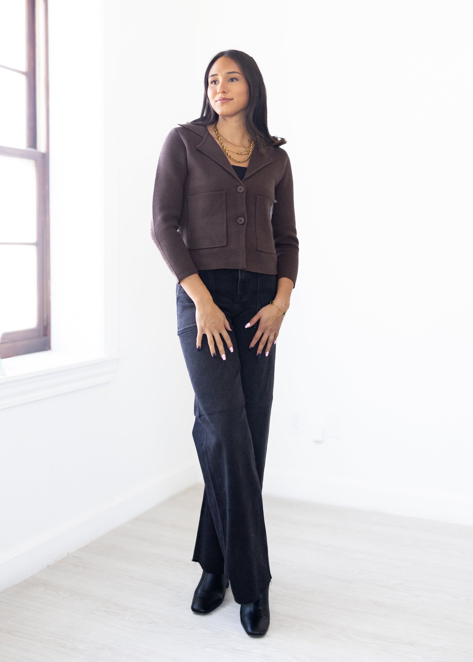 Woman wearing a brown jacket and dark jeans standing in a bright room.