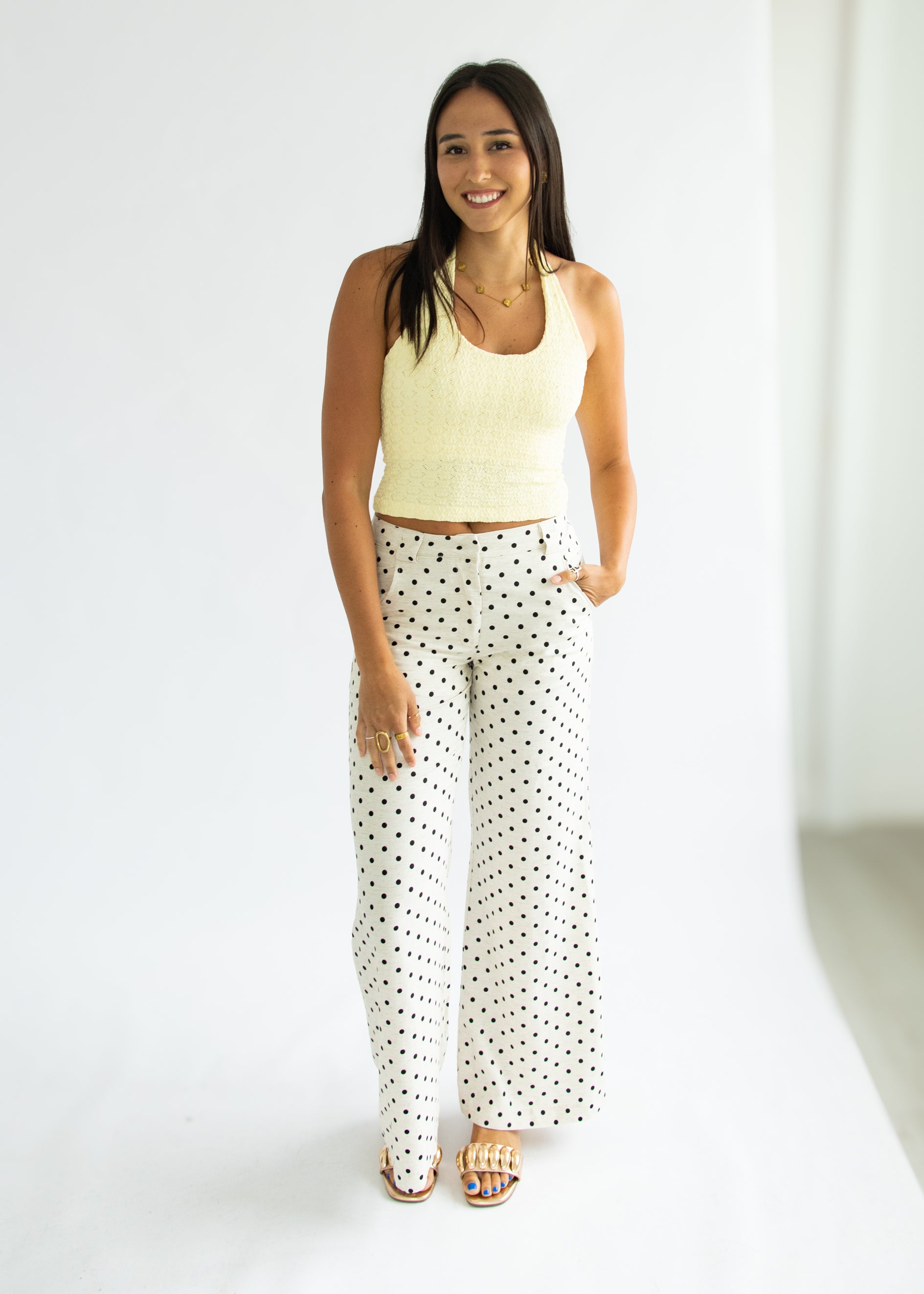 Woman wearing a light yellow sleeveless top and white polka dot pants on a white background