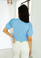 Person wearing a light blue blouse with puffed sleeves and white pants, standing against a white wall.