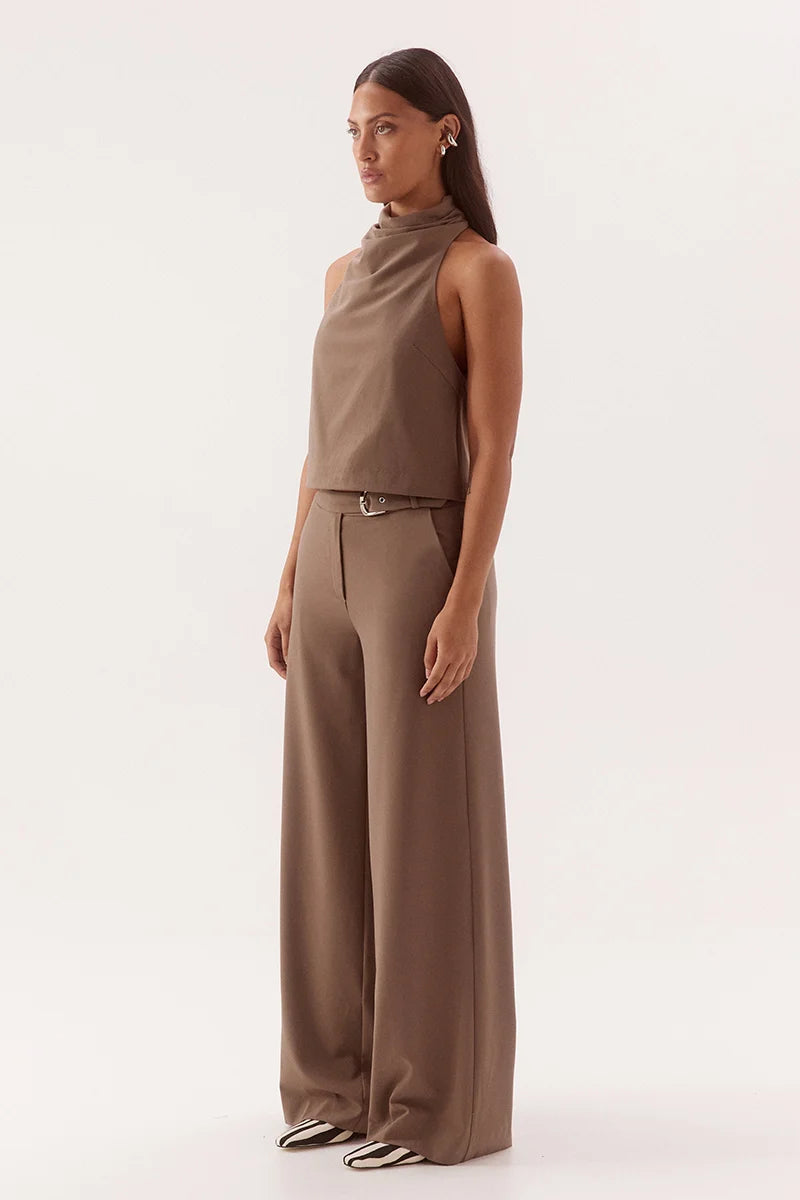 A woman modeling a taupe-brown sleeveless high-neck top and matching wide-leg trousers with a thin belt, photographed against a clean off-white studio background.
