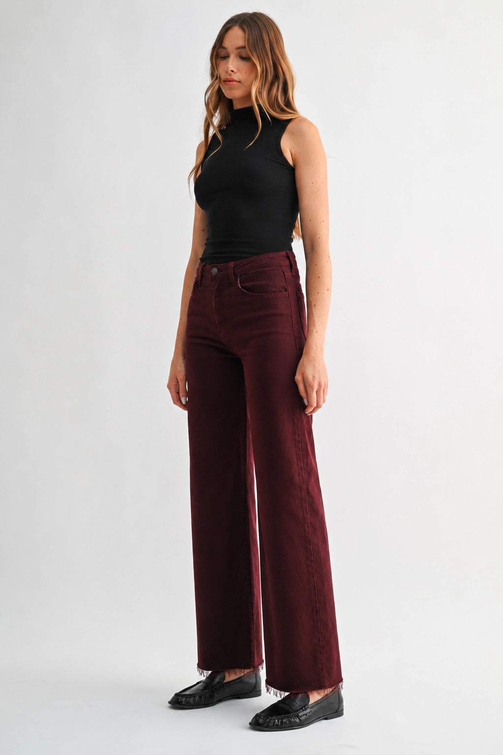 A woman modeling a pair of high-waisted, wide-leg trousers in a deep burgundy velvet fabric, paired with a black sleeveless top and black loafers, against a plain light gray studio background.