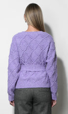 A woman viewed from the back wearing a lavender purple textured knit sweater with a waist tie and gray trousers, standing against a plain light gray wall.