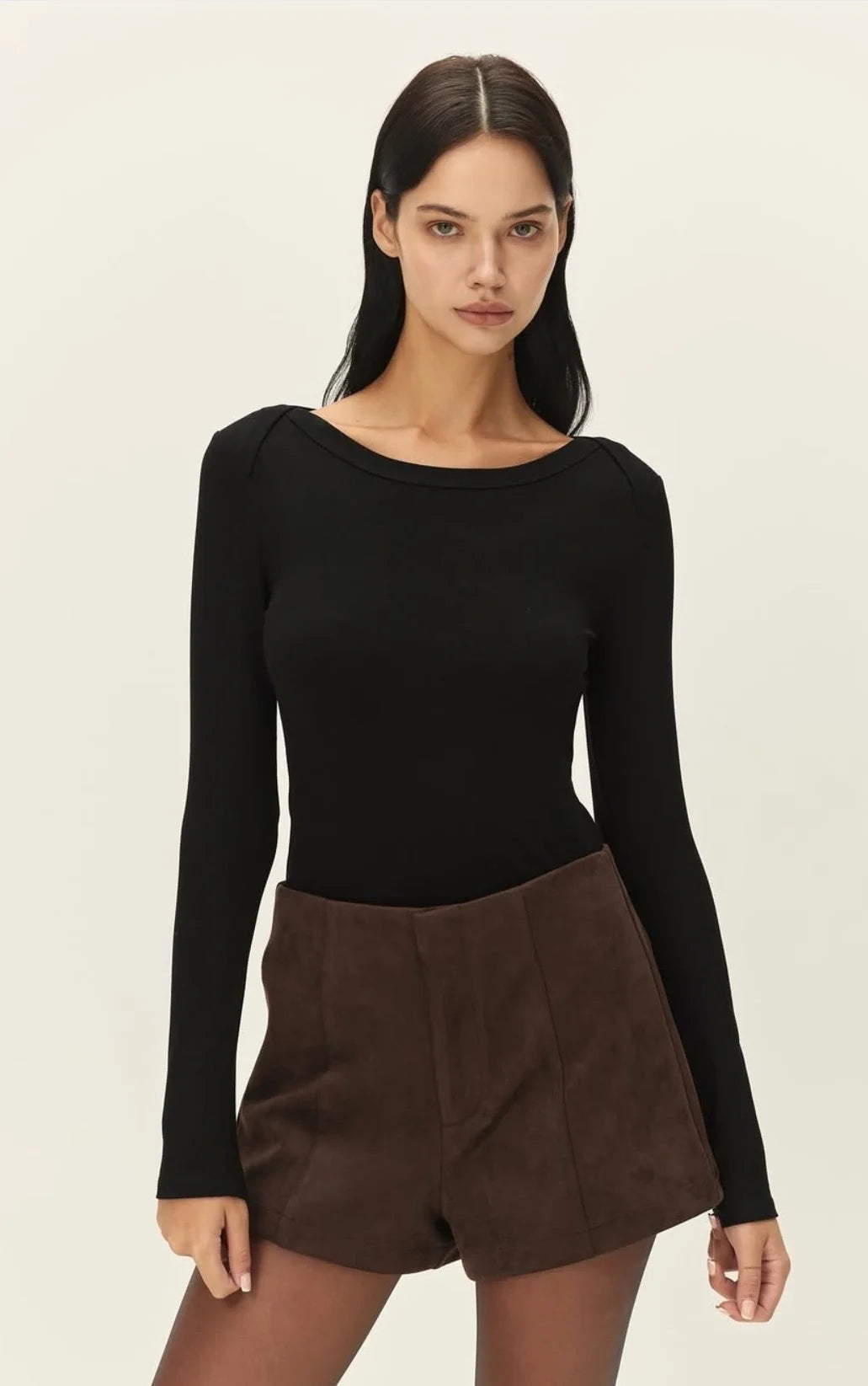 A woman modeling a black long-sleeve boat-neck top paired with brown shorts against a plain light beige studio background.