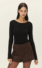 A woman modeling a black long-sleeve boat-neck top paired with brown shorts against a plain light beige studio background.