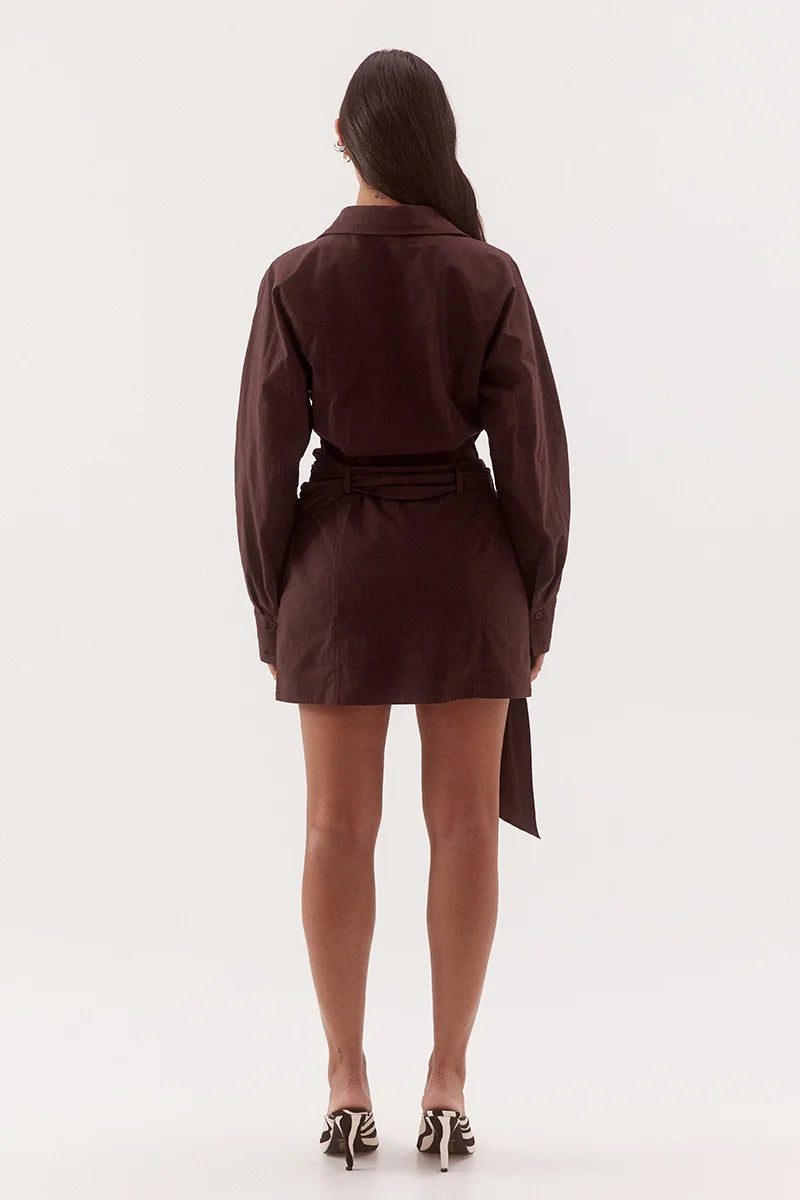 A woman viewed from the back wearing a dark brown long-sleeve mini wrap dress with a tie waist, paired with black and white patterned heels, against a plain white studio background.