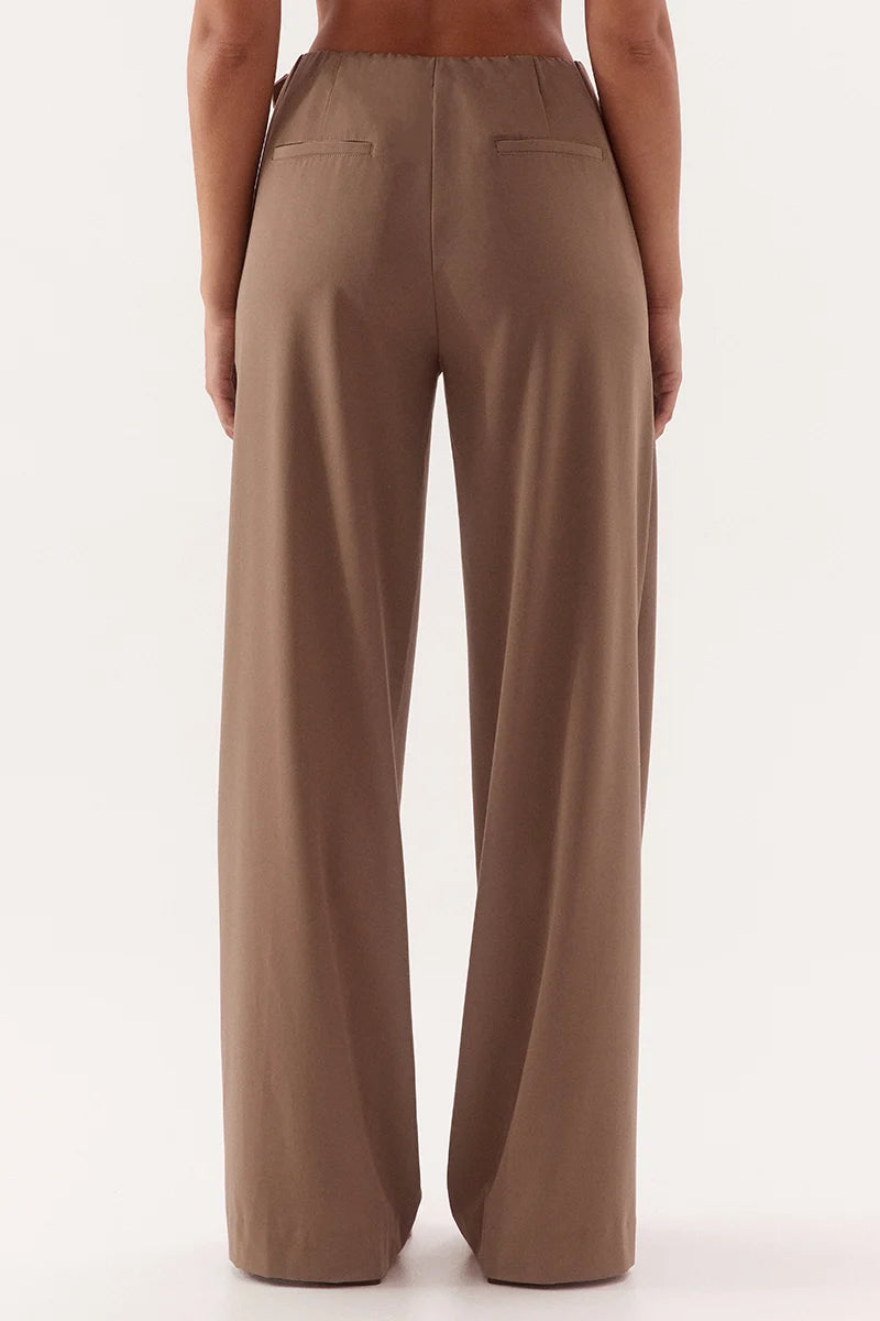 A person shown from the back wearing high-waisted, wide-leg trousers in a warm taupe-brown color, photographed against a clean off-white studio background.