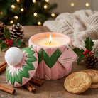 A pink ceramic candle jar with a matching lid sits on a wooden table, lit and surrounded by holiday props including pinecones, holly, cinnamon sticks, cookies, and a knitted blanket. The lid has a white pom-pom and green-and-pink chevron detailing.