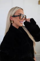Woman wearing a black fur coat and large sunglasses indoors.