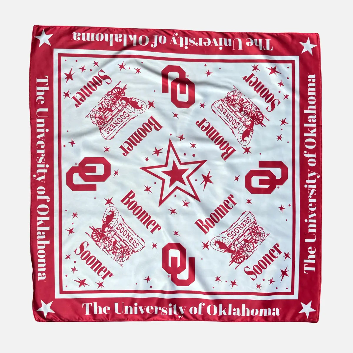 A square University of Oklahoma themed bandana or scarf laid flat on a light gray background. The design features a white center with red logos, stars, and text, surrounded by a red border with white stars and 'The University of Oklahoma' printed along the edges.