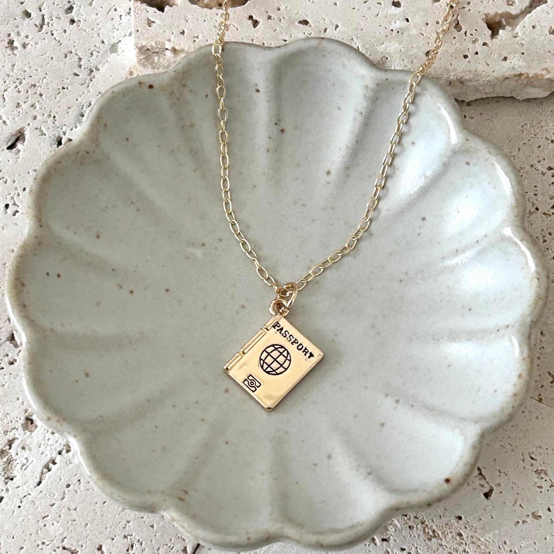 A gold necklace with a miniature passport book pendant displayed on a scalloped ceramic dish over a textured stone surface.
