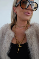 Person wearing a gold chain necklace with a cross pendant, black sunglasses, and a fur coat.