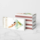 A novelty matchbox set with a retro illustration of a woman in a red bikini drinking from a martini glass filled with olives. One matchbox is open, showing white matches inside, and another stack of three closed matchboxes sits behind it on a light marble surface against a white background.