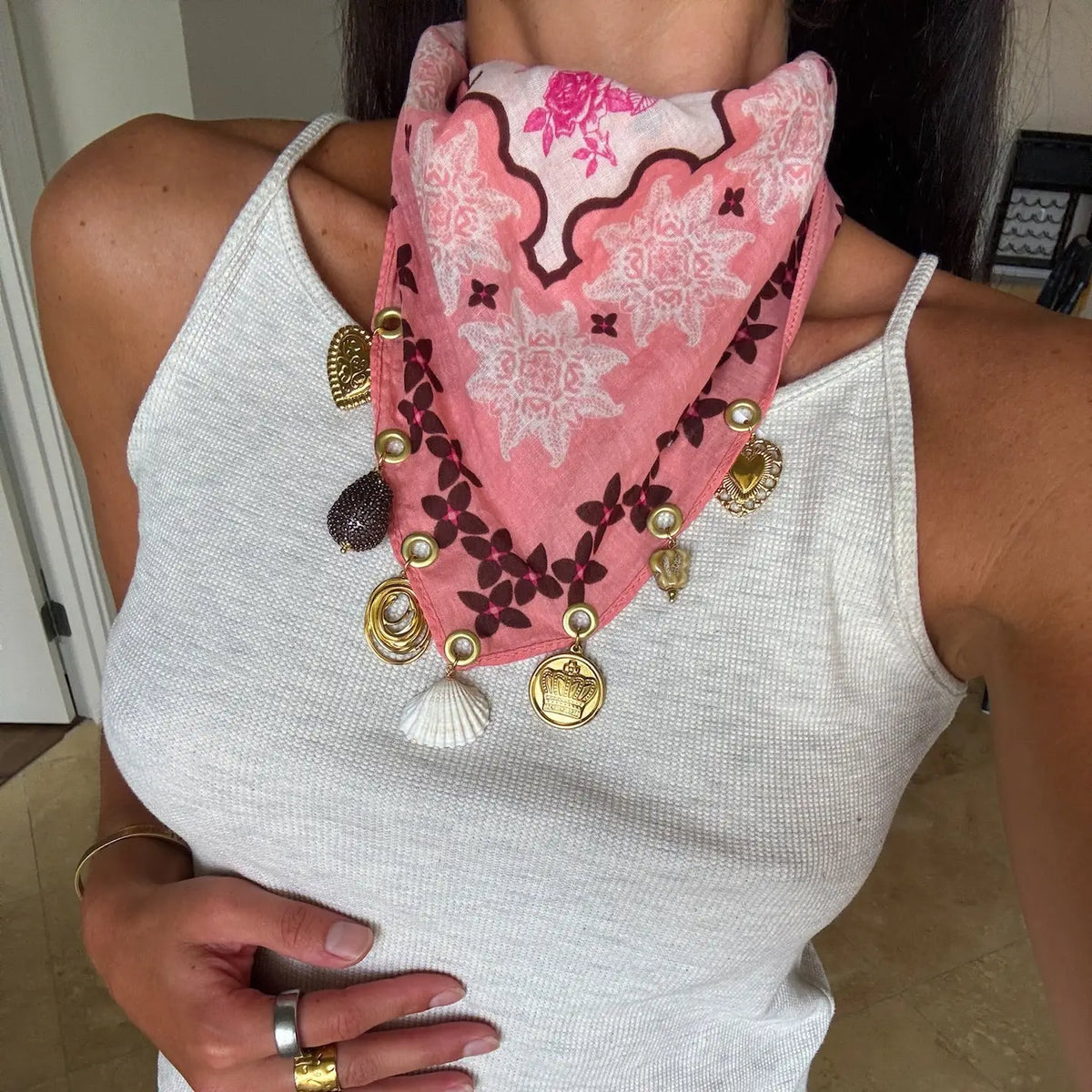 A pink patterned bandana styled as a bohemian necklace with gold coins, shells, and beads, worn by a person in a white ribbed tank top.